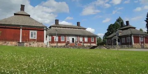 Malingsbo herrgård med sina röda trähus och stenfundament, omgivet av en grönskande äng med vita blommor under en blå himmel med moln.