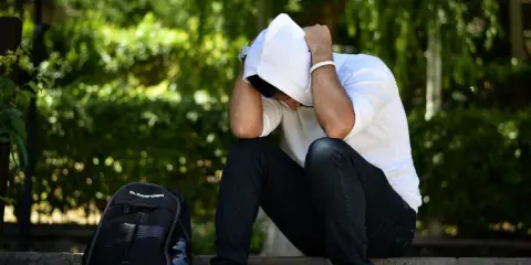 Młody mężczyzna w białej bluzie z kapturem siedzi ze spuszczoną głową, ręce na twarzy. Obok plecak. Czy to stres, a może wysoki kortyzol? Co to jest kortyzol?
