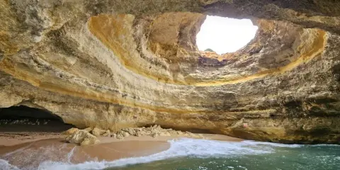 Die Benagil-Höhle mit ihrem kreisförmigen Loch im Felsdach, das Sonnenlicht hereinlässt. Wellen brechen sanft am Sandstrand im Inneren.
