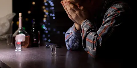 Mężczyzna z rękami na twarzy przy barze, obok butelka i kieliszek. Obraz symbolizuje przymusowe leczenie alkoholika i jego walkę.