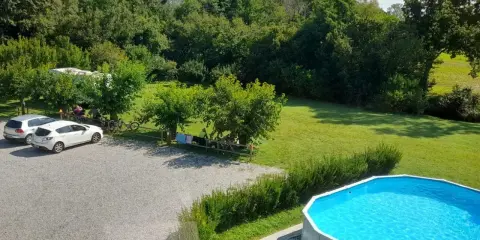 Un **agricampeggio con piscina** immerso nel verde. Auto parcheggiate, camper, biciclette e persone che si rilassano all'ombra degli alberi. La piscina azzurra invita a un tuffo rinfrescante.