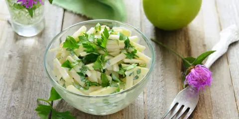 Frischer Kohlrabi Salat mit Petersilie in einer Glasschüssel, garniert mit einer lila Blume und grünen Äpfeln auf Holztisch.
