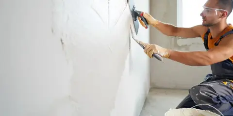 Un ouvrier, protégé par des lunettes et des gants, s'applique à crépire un mur avec une truelle.
