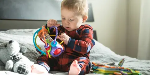 Małe dziecko w kratkę odkrywa świat, kiedy niemowlę zaczyna chwytać kolorową grzechotkę. Obok leży pluszowy miś.