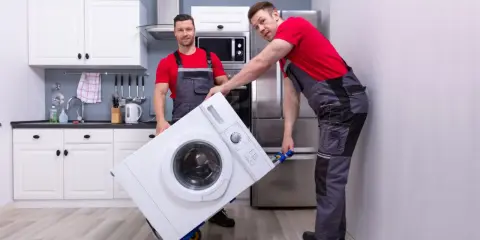 Dwóch mężczyzn w roboczych ubraniach przewozi pralkę na wózku. Czy można przewozić pralkę na leżąco? To pytanie zadaje sobie wielu przy przeprowadzce.