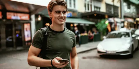 Ung man med mobiltelefon i handen, på en stadsgata med bilar och butiker. Känns som vårt hem utomlands.