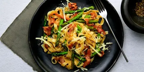 Pyszny makaron ze szparagami, chrupiącym boczkiem i parmezanem na czarnym talerzu. Idealny na wiosenny obiad.