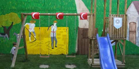 Un niño observa un columpio y un tobogán azul, parte de unos juegos medievales para niños, con un mural de fondo.