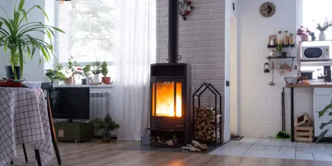 Płonący kominek z podłączeniem do komina, obok stos drewna. W tle kuchnia i okno z firanką.