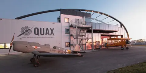 Zwei historische Flugzeuge stehen vor dem Quax Hangar. Ein graues Propellerflugzeug im Vordergrund, ein gelbes Doppeldeckerflugzeug daneben.