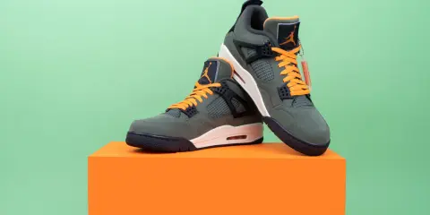 Jordan Motorsport Sneaker in Olivgrün mit orangefarbenen Schnürsenkeln, auf einem orangefarbenen Podest vor hellgrünem Hintergrund.
