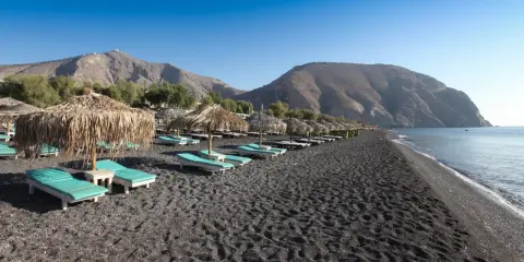 Santorini spiagge di sabbia nera con lettini e ombrelloni di paglia, montagne sullo sfondo e mare calmo.