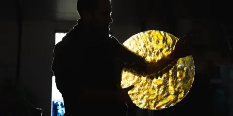 Artisan tenant un disque de verre texturé, illuminé par derrière. Les reflets dorés rappellent des vitraux.