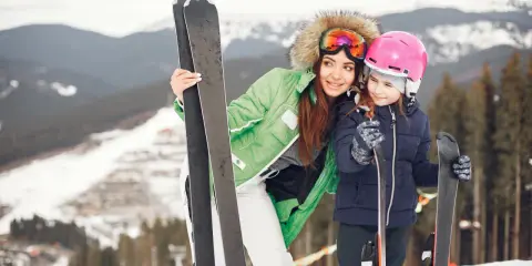 Kobieta i dziecko w strojach narciarskich z nartami na tle górskiego stoku.
