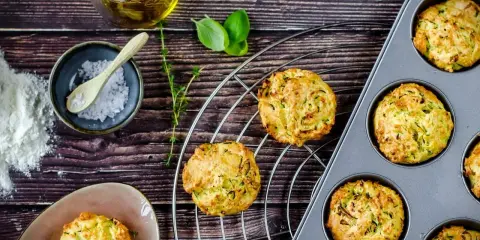 Goldbraune Zucchini-Muffins auf einem Gitter, einige in einer Muffinform. Zutaten wie Öl, Salz und Kräuter liegen bereit.