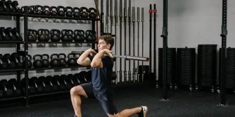 Hombre realizando un front rack lunge con barra en un gimnasio, rodeado de pesas y barras.