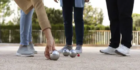 Une main de nom joueur de pétanque français saisit une boule argentée sur un terrain de gravier. D'autres boules et un cochonnet sont visibles.