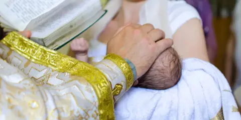 Ksiądz w złotej szacie trzyma dziecko do chrztu, czytając z księgi.