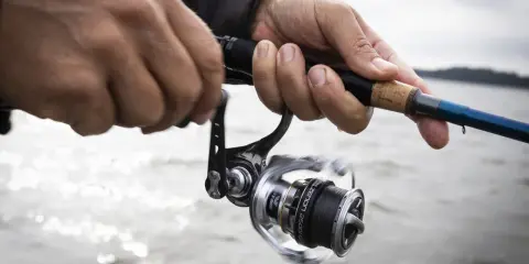 Händer som håller i ett fiskespö med en rulle, redo för fiska i Sverige. Vattnet glittrar i bakgrunden.