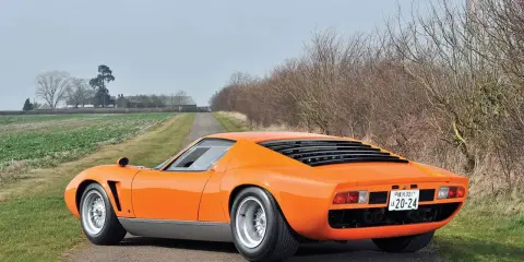 Ein leuchtend oranger Lamborghini Jota parkt auf einer einsamen Landstraße, umgeben von Feldern und kahlen Bäumen.
