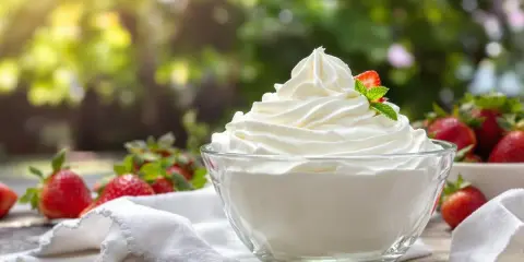Un bol de crème chantilly qui tient longtemps, garni d'une fraise et d'une feuille de menthe, entouré de fraises fraîches.