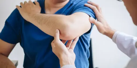 Fizjoterapeuta bada łokieć pacjenta. Dłonie specjalisty delikatnie dotykają bolącego miejsca, oceniając zakres ruchu i ból.