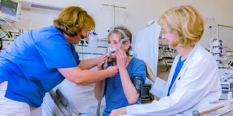 Ärztin und Pflegekraft helfen Patientin mit Lungenemphysem bei der Anpassung einer Beatmungsmaske. Medikamente sind Teil der Behandlung.