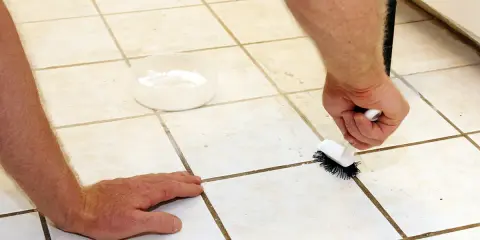 Mann reinigt mit Bürste und einer Schale mit weißer Paste die Fliesenfugen, um sie zu erneuern.