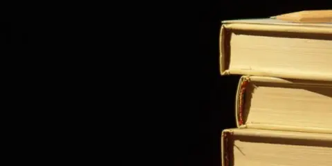 Libri impilati, un libro in cima con una matita. La luce crea ombre, un po' come l'anastrofe figura retorica che inverte l'ordine per enfasi.