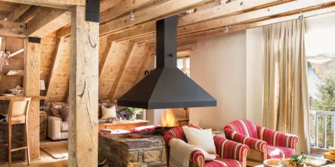 Chimenea central de diseño moderno, uno de los tipos de chimeneas que aporta calidez a este salón rústico con vigas de madera.