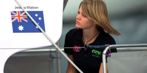 Jessica Watson heute, blickt nachdenklich auf dem Boot. Die australische Flagge ist im Hintergrund zu sehen.