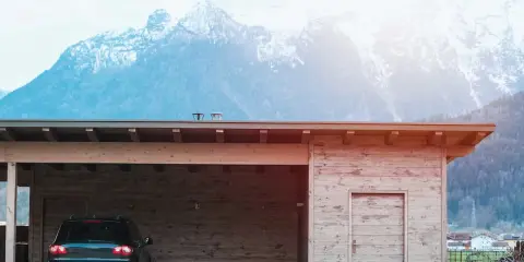 En nybyggd carport i trä med en bil parkerad under. Bakom syns snöklädda berg.