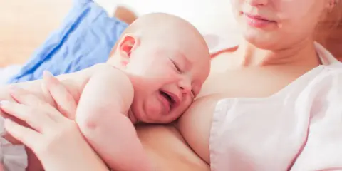 Ein Baby schreit beim Stillen, die Augen geschlossen, das Gesicht verzogen. Die Mutter hält es sanft.