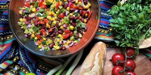 Bunter mexikanischer Salat mit Mais, Bohnen und Gemüse in einer Schüssel, daneben Baguette, Tomaten und Kräuter.