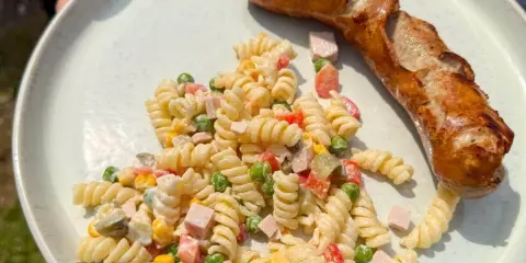 Gegrillte Wurst mit einem cremigen Nudelsalat als Beilage, garniert mit Erbsen, Mais und gewürfeltem Schinken.