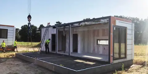 Budowa domu na zgłoszenie postępuje. Dźwig podnosi moduł, a pracownicy w kamizelkach nadzorują montaż. Warunki zabudowy sprzyjają szybkiemu postawieniu konstrukcji.