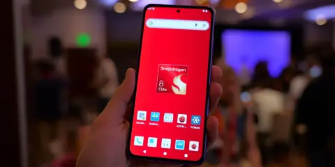 Dłoń trzyma smartfon z czerwonym tłem i ikoną Snapdragon 8 Elite. Widoczne ikony benchmarków, sugerujące testowanie wydajności procesora.