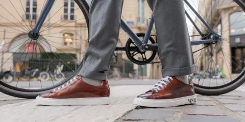 Eleganckie sneakersy do garnituru w brązowym kolorze, zestawione z szarymi spodniami, na tle miejskiego krajobrazu z rowerem.