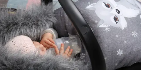 Noworodek w kombinezonie z futerkiem i czapką trzyma butelkę z mlekiem w foteliku samochodowym.
