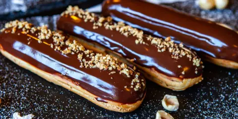 Trois éclairs au chocolat brillants, saupoudrés de noisettes concassées, sur un fond sombre parsemé de sucre glace.
