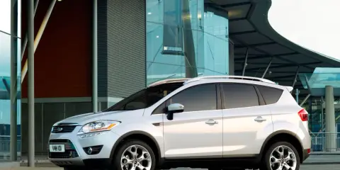 Biały Ford Kuga 2010 zaparkowany przed nowoczesnym budynkiem z przeszkloną fasadą.