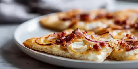 Tranches de flammekueche recette originale sur une assiette blanche, garnie de lardons, oignons rouges et fromage crémeux.