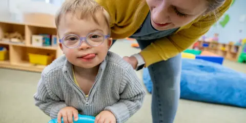 Chłopiec z zespołem Downa, w okularach, z językiem na brodzie, bawi się z terapeutką. To przykład pracy w obszarze oligofrenopedagogiki.