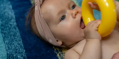 Niemowlę w opasce na głowie wkłada do buzi żółtą zabawkę na tle kolorowego koca.