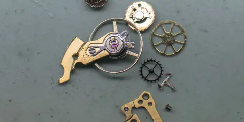 Parti di un orologio: ingranaggi, un bilanciere e altri componenti metallici disposti su una superficie grigia.