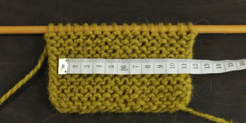 Knitted swatch with a measuring tape across it, showing stitches on wooden needles.