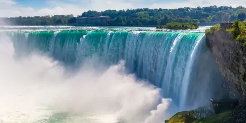 Potężny wodospad Niagara w całej okazałości. Woda spada kaskadami, tworząc mgłę. W tle drzewa i budynek.