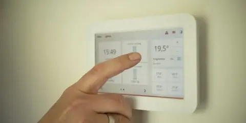 Dłoń z pierścionkiem na palcu dotyka ekranu termostatu, być może regulując ustawienia, aby uniknąć częstego włączania i wyłączania pieca gazowego.