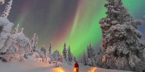 Un uomo con lanterna, vestito da Babbo Natale, ammira l'aurora boreale in Lapponia. Un paesaggio innevato e magico, perfetto per chi cerca dove andare in Lapponia.