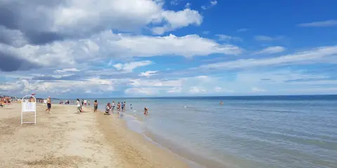 Una giornata di sole a Riccione mare, con gente che passeggia sulla sabbia e si gode l'acqua cristallina sotto un cielo azzurro con nuvole bianche.